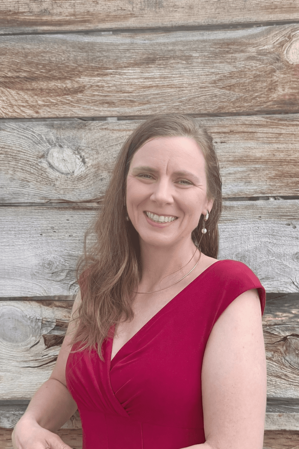 Smiling woman with long brown hair wearing a red dress against weathered wooden planks.